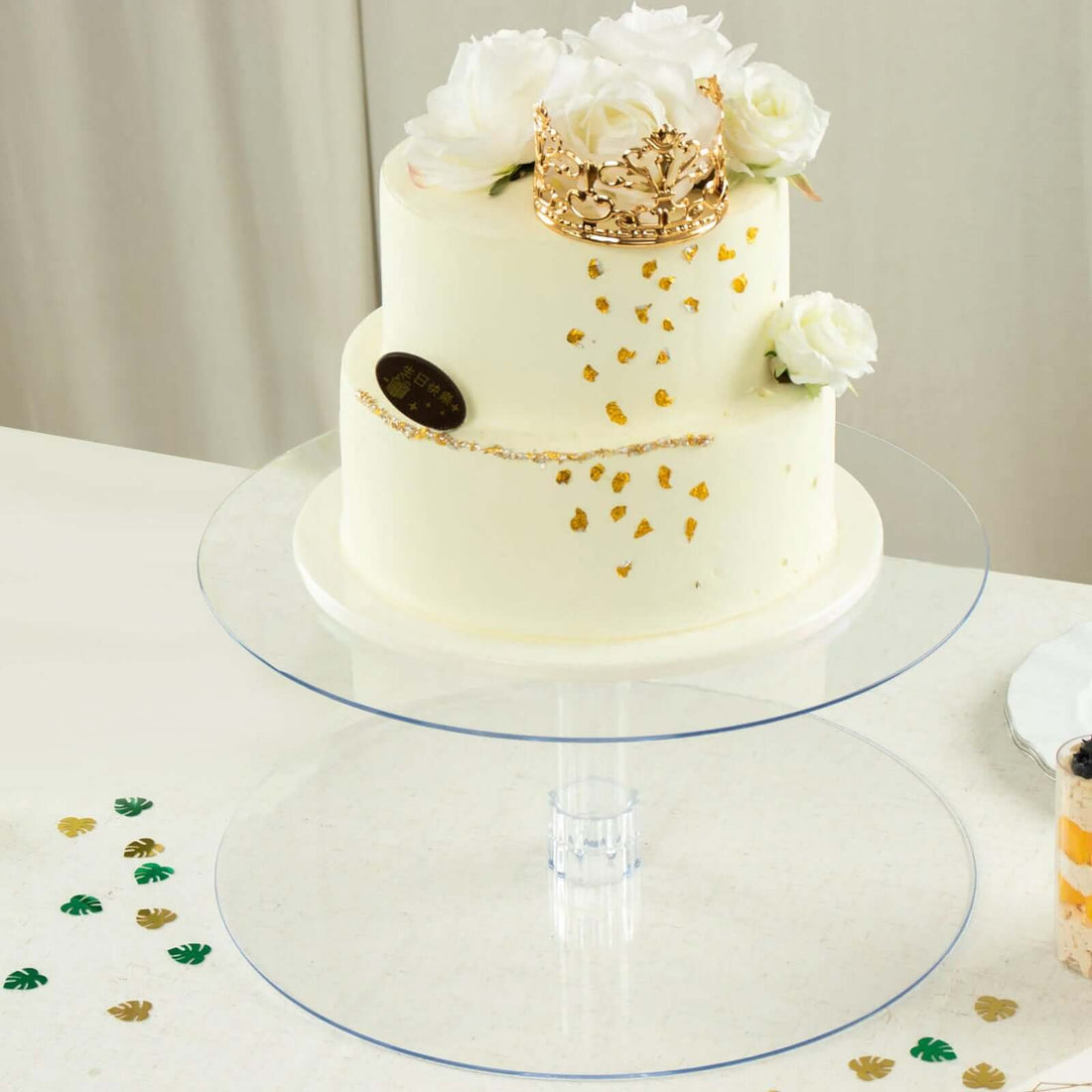 Elegant white cake adorned with roses and a gold crown on a clear cake stand.