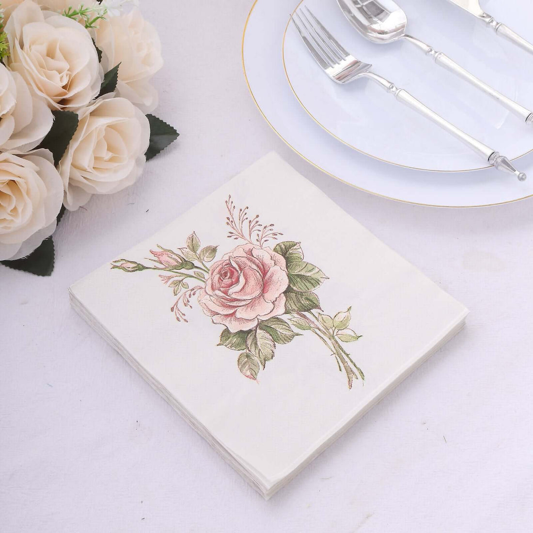 Floral design paper napkin on a table setting with silver cutlery and roses.