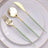 Gold and green utensils set on a white plate with golden rim.