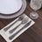 Elegant gray cutlery set on a textured napkin with a plate and glass in a rustic setting.