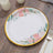 Floral design paper plate with gold rim on wooden surface.