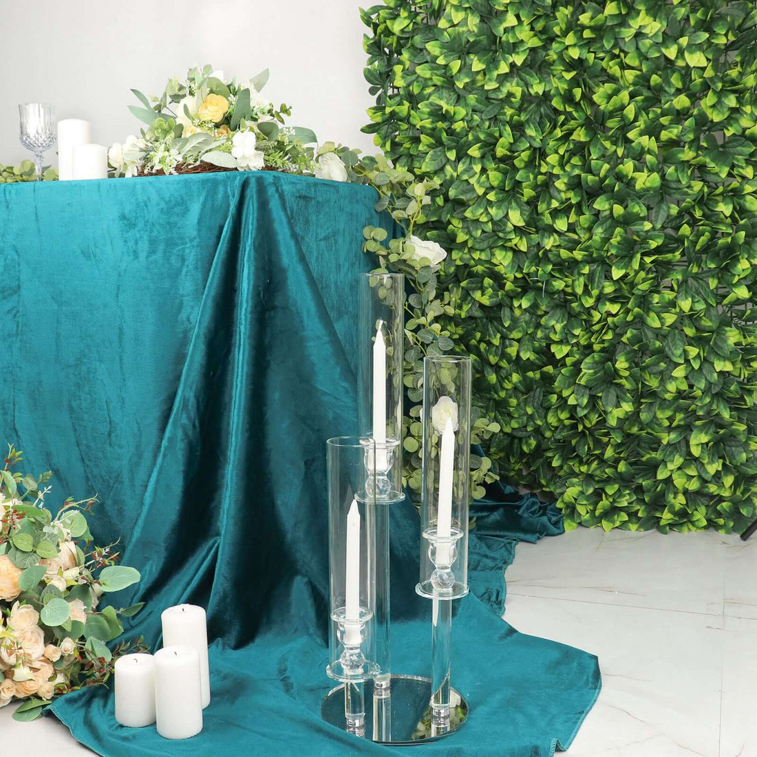 Elegant glass candle holders on a teal table with greenery backdrop.