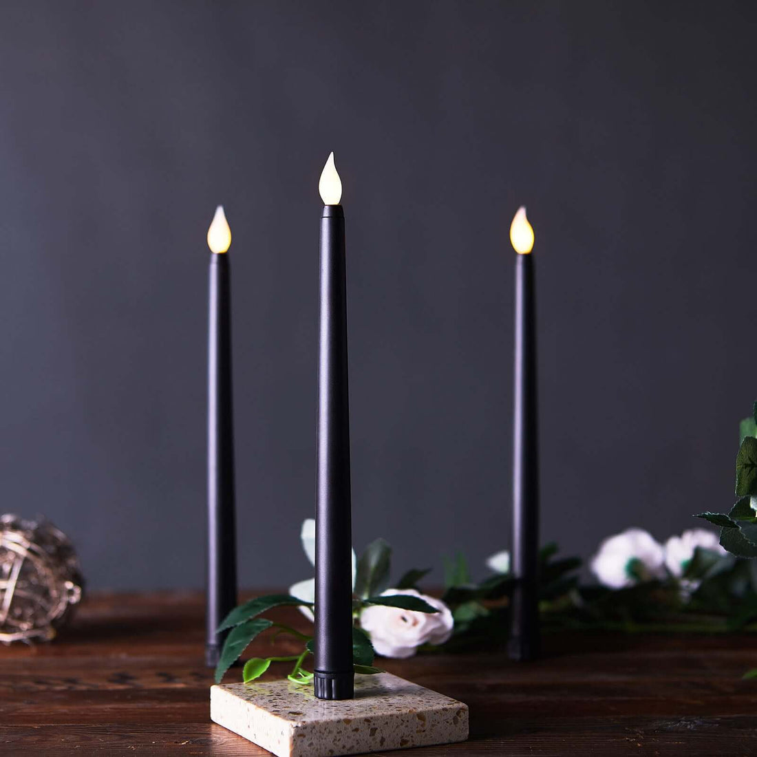 Black LED taper candles on a stone base with greenery and flowers.