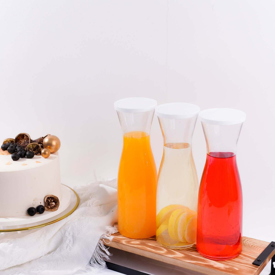 Three glass carafes with colorful drinks on a wooden board beside a cake.