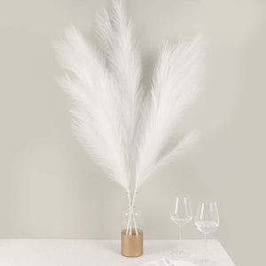 White pampas grass in a gold vase with wine glasses on a table.