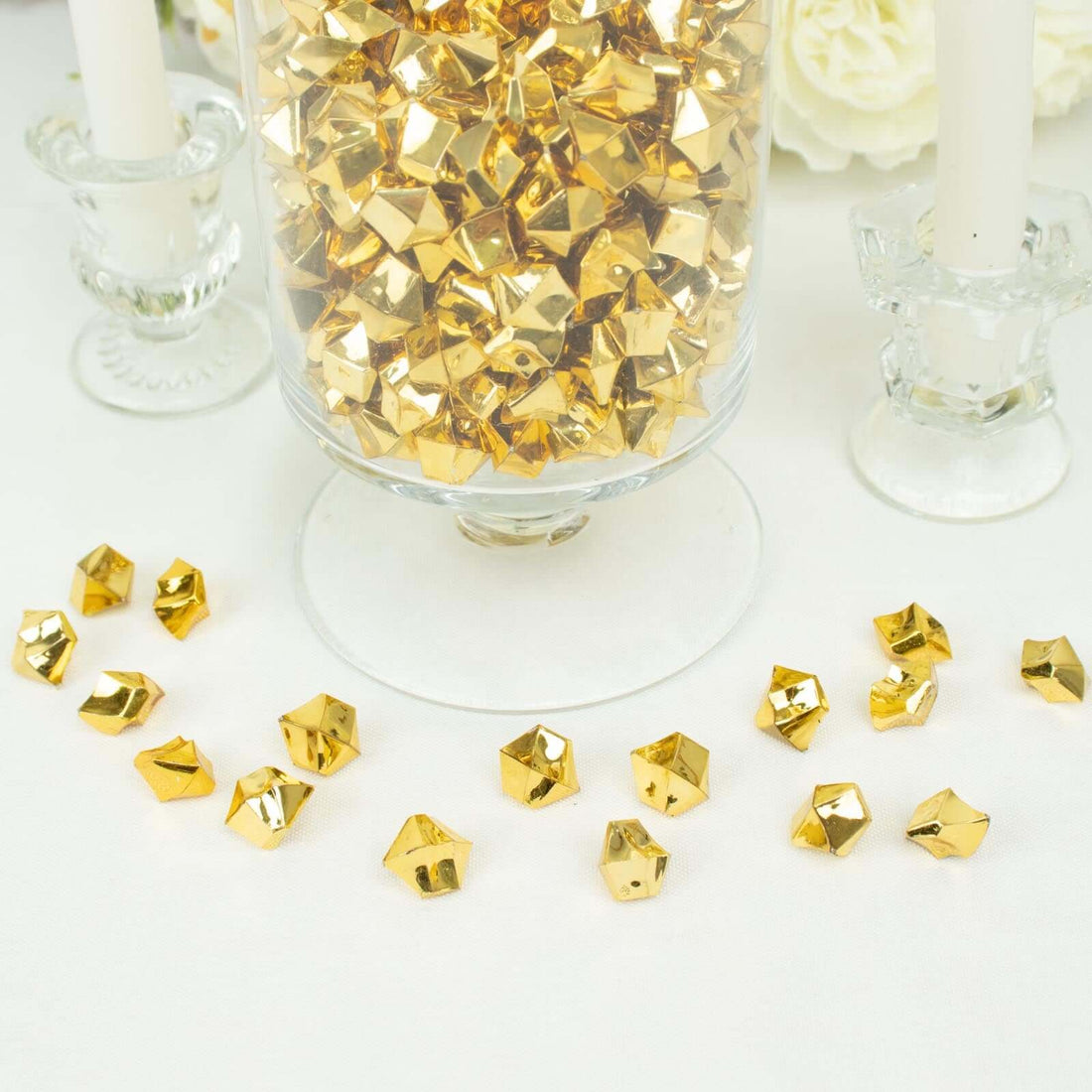 Gold decorative gems in a glass jar with scattered pieces on a soft surface.