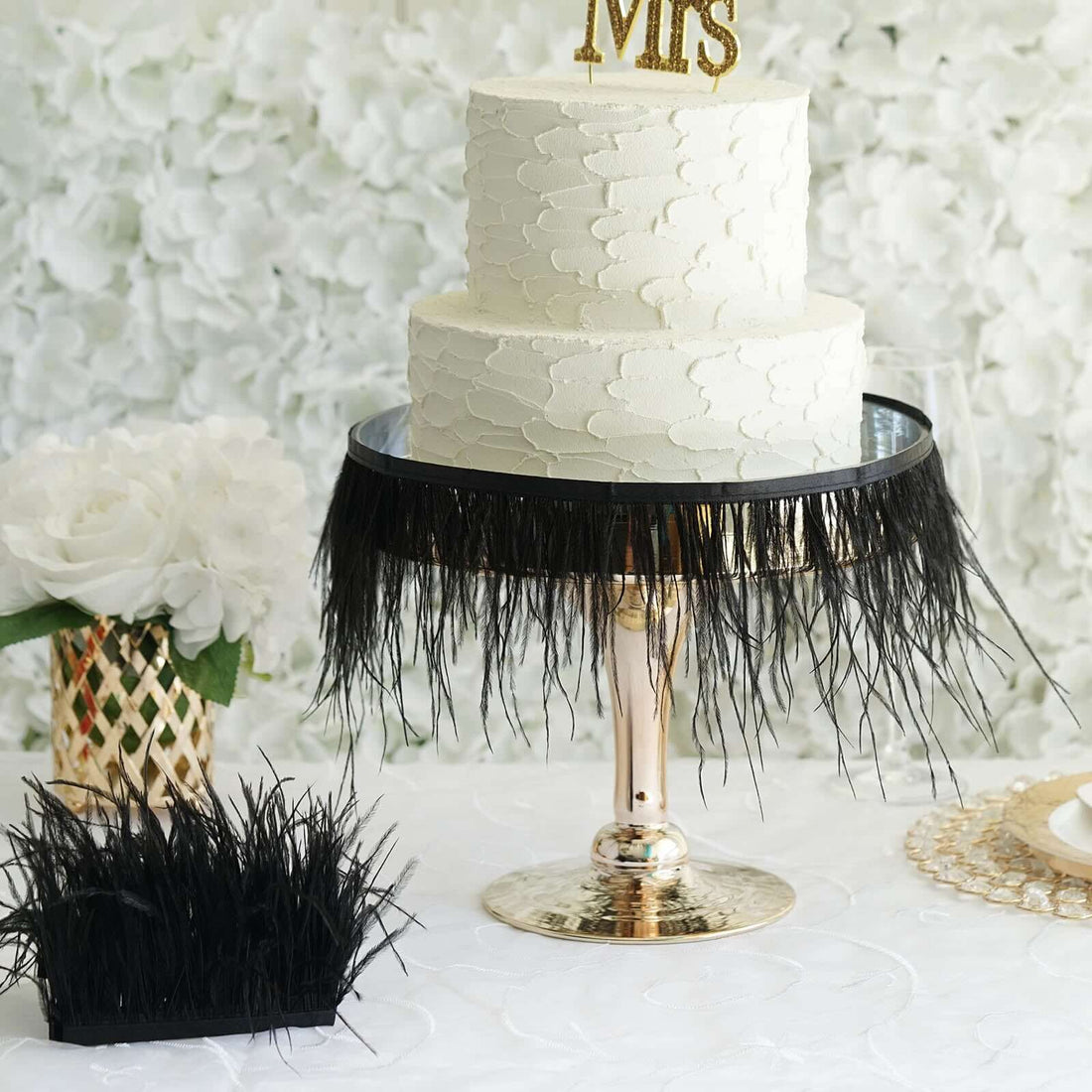 Elegant wedding cake with textured frosting and decorative black fringe, set against floral backdrop.