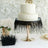 Elegant wedding cake with textured frosting and decorative black fringe, set against floral backdrop.