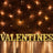 "Illuminated 'VALENTINES' letters against a brick wall with string lights."