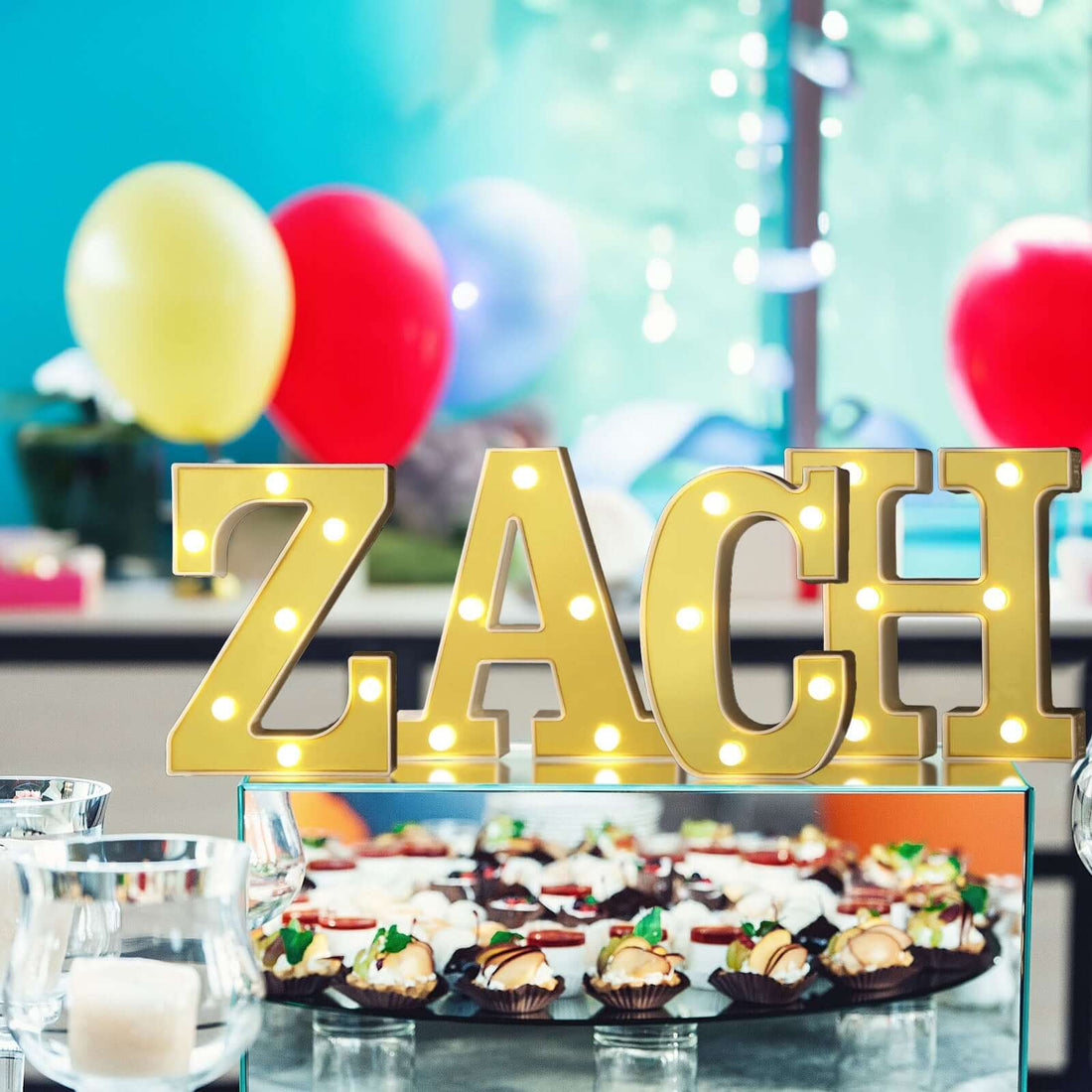 Gold marquee letters "ZACH" with colorful balloons and desserts in the background.