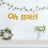 "Oh Baby" banner with greenery and floral arrangement in a staged decor setting.