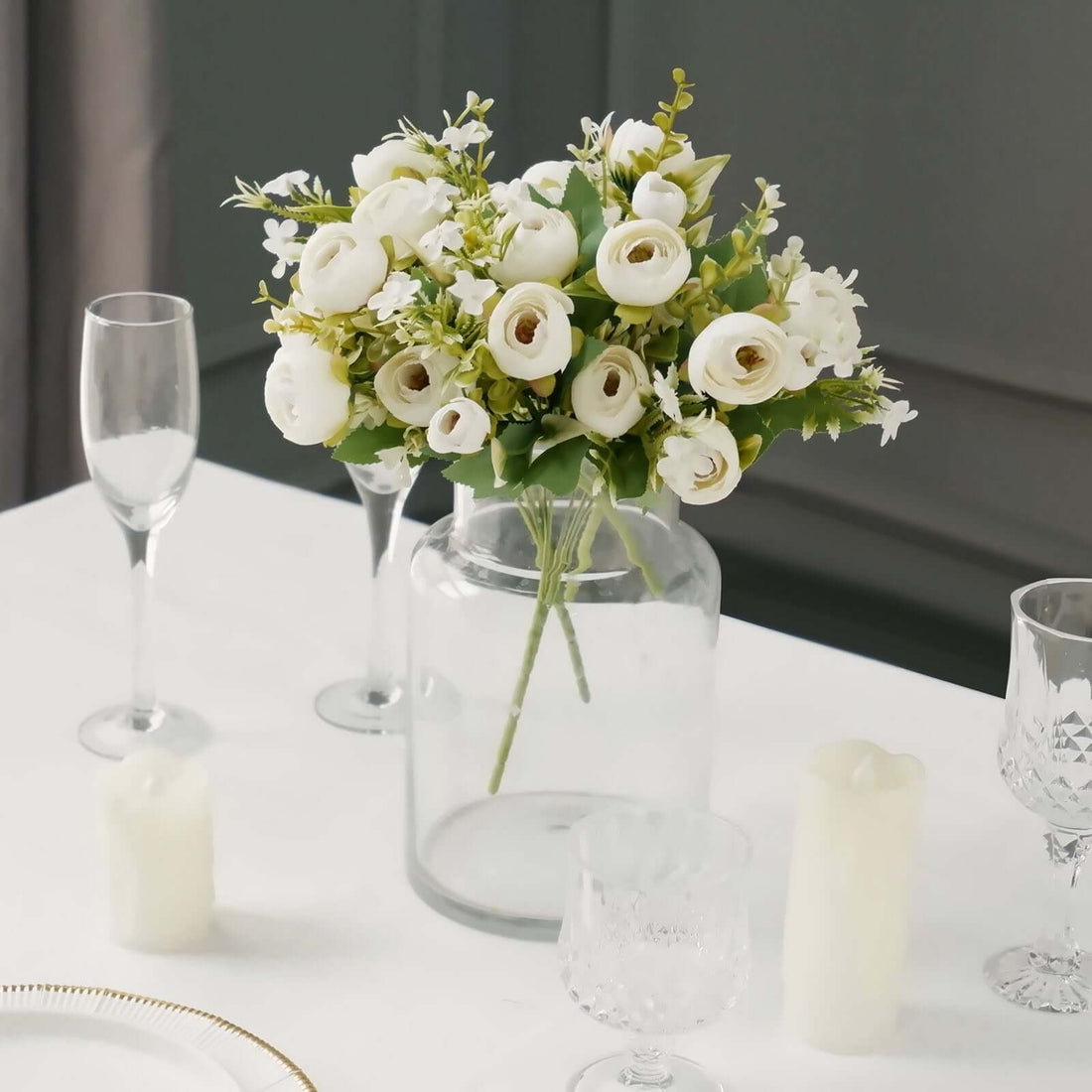 White flower bouquet in a glass vase on a dining table with candles and glasses.