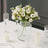 White flower bouquet in a glass vase on a dining table with candles and glasses.