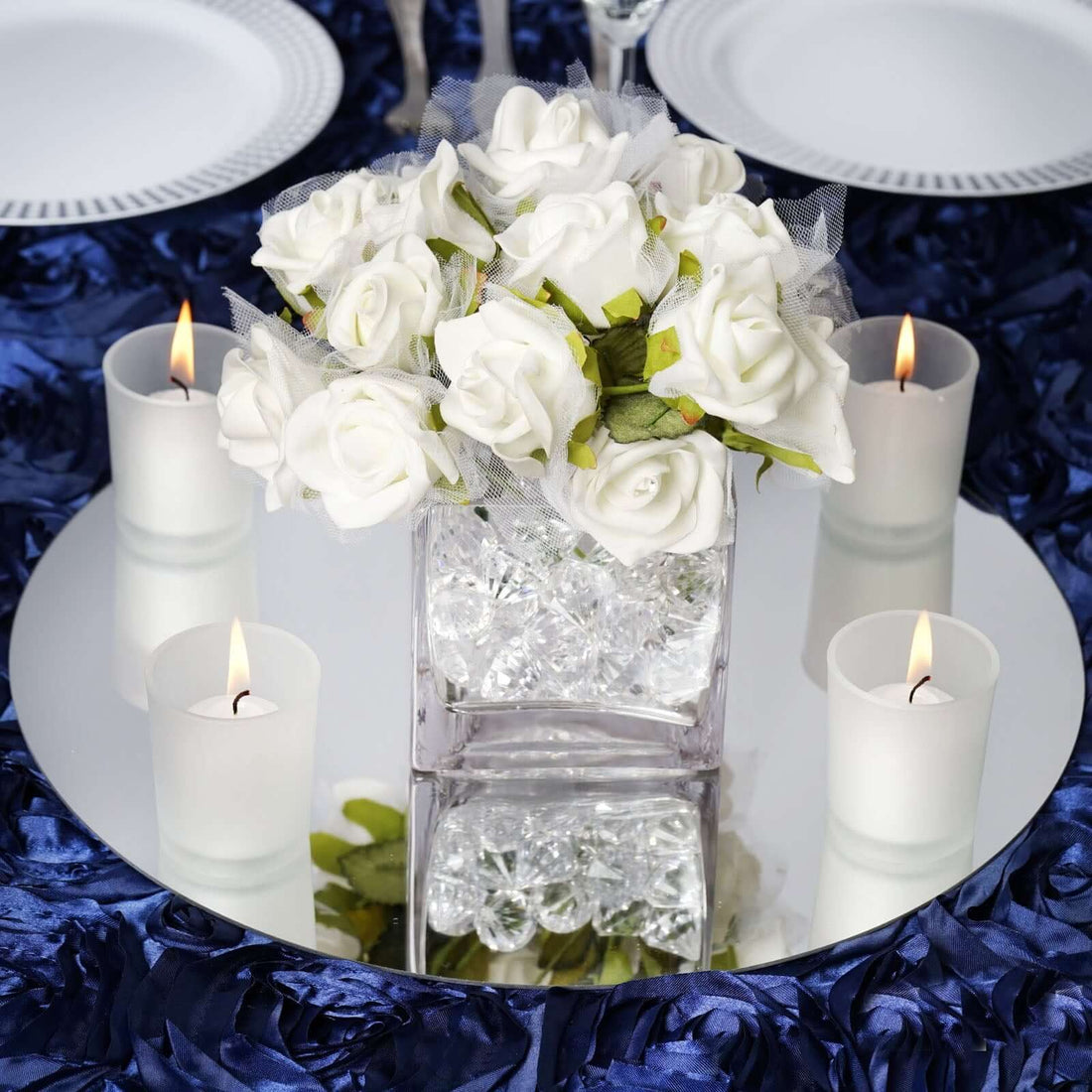 Elegant white rose centerpiece with candles on a reflective surface.