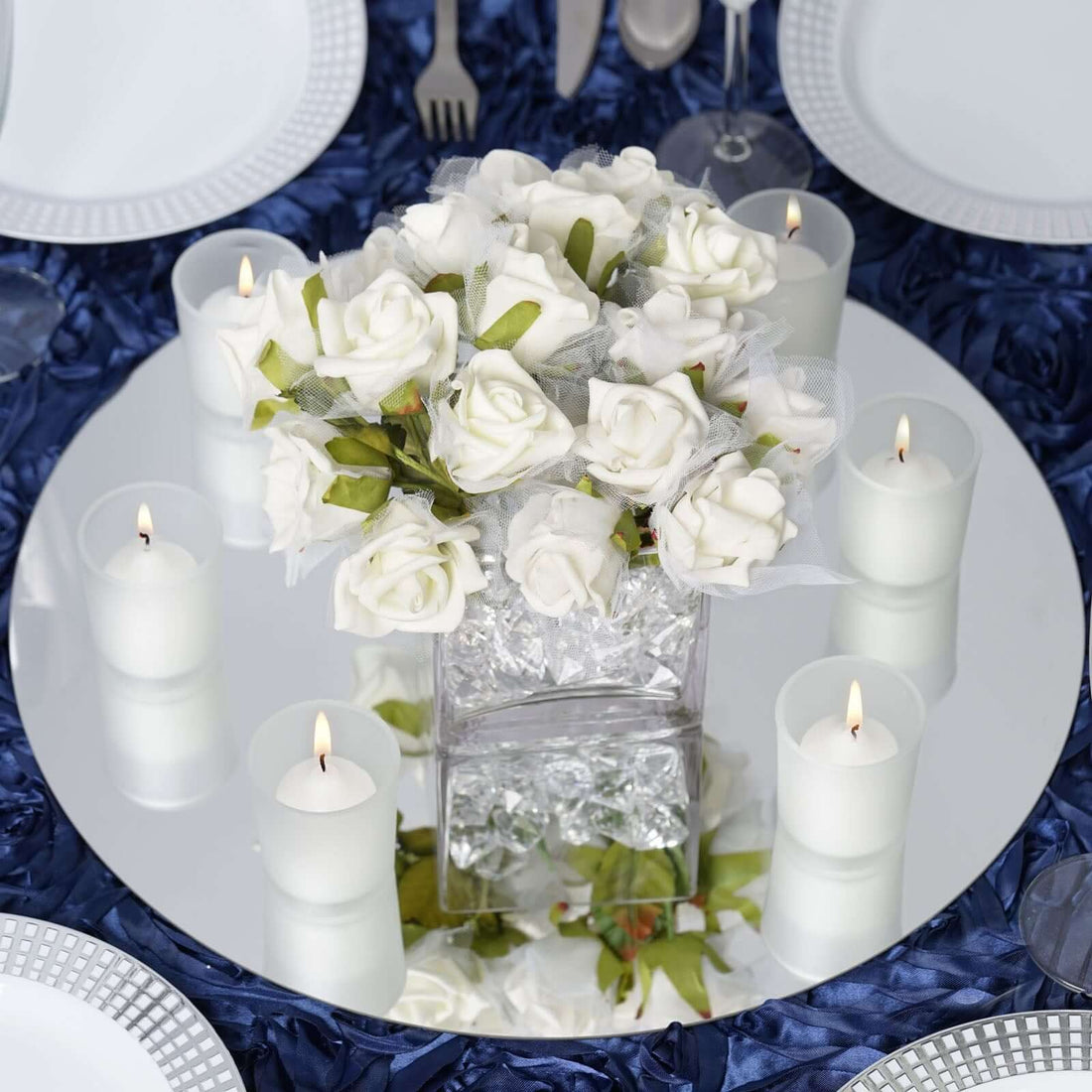 White rose bouquet centerpiece with candles on a mirrored table setting.