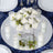 White rose bouquet centerpiece with candles on a mirrored table setting.