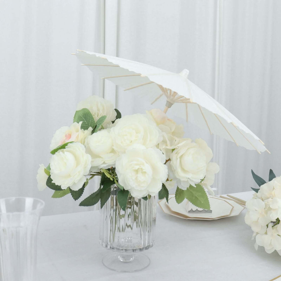 White roses in a glass vase with a white parasol on a table.