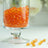 Orange crystal gems in a glass jar with scattered pieces on a white surface.