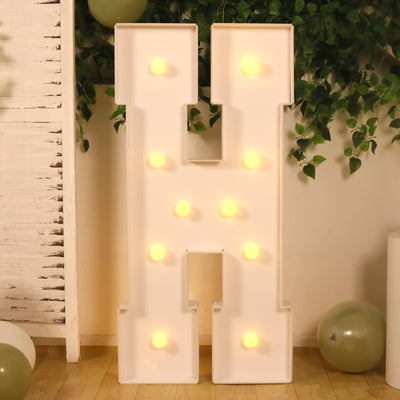 White illuminated letter H marquee sign with warm bulbs and greenery background