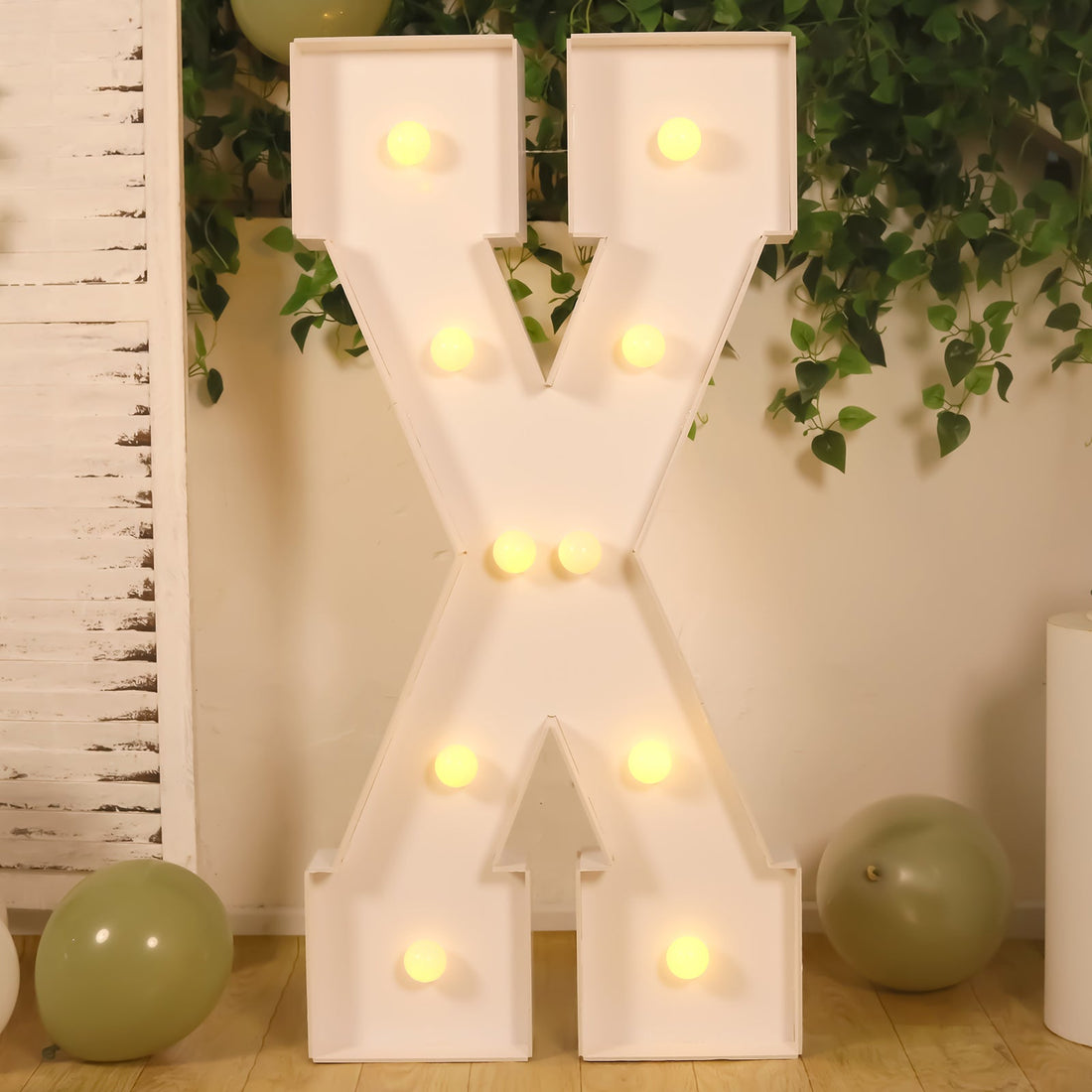 Light-up marquee letter "X" in white with bulbs, set against greenery and balloons.