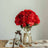 Red artificial flowers in a glass vase with a ribbon on a wooden table.