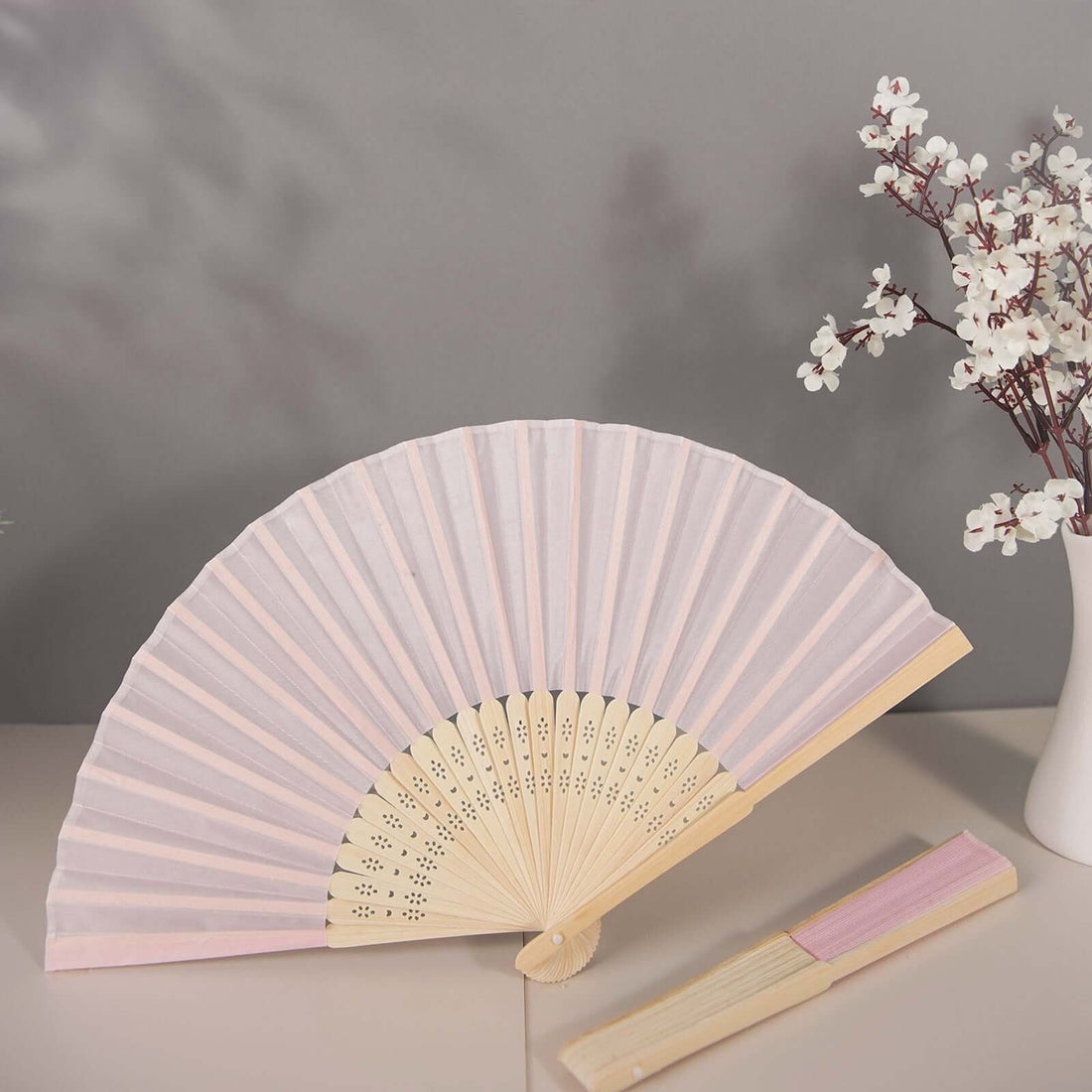 Pink folding fan with wooden handle and floral decor, displayed on a table.