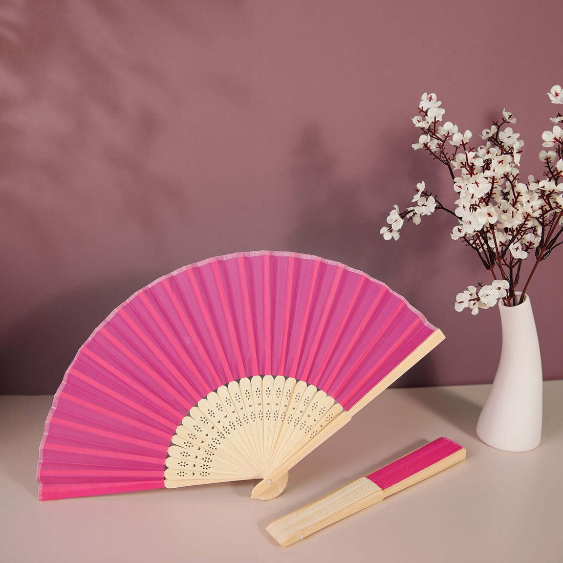 Pink folding fan with cream accents next to a vase of white flowers.