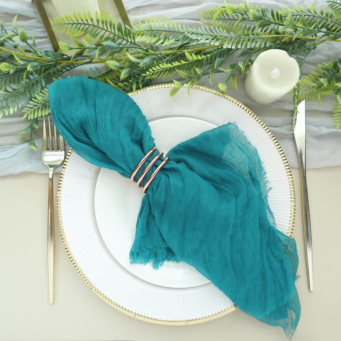 Teal napkin with gold rings on a white plate, surrounded by greenery and candles.