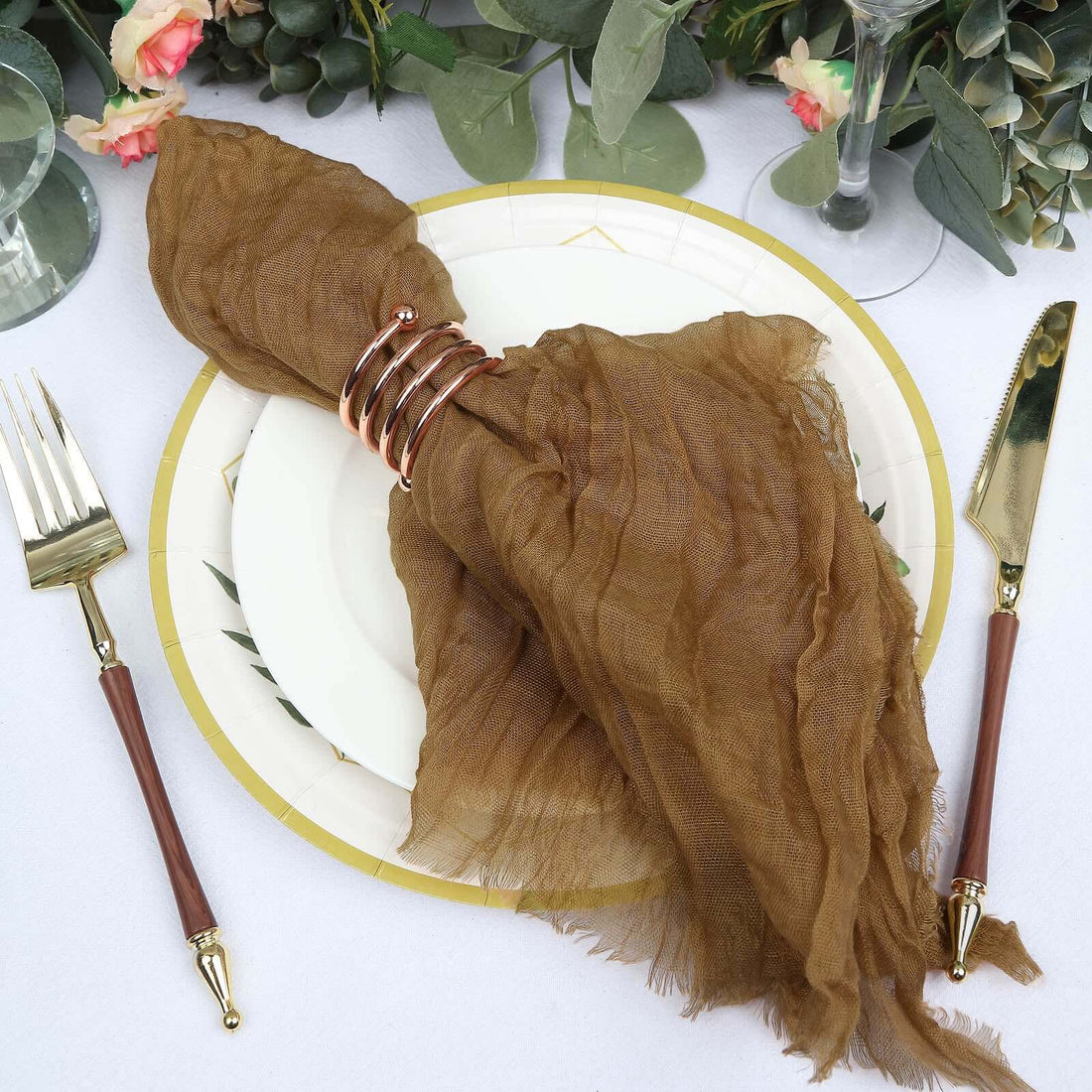 Brown textured napkin with a gold ring on a white plate, surrounded by elegant tableware.