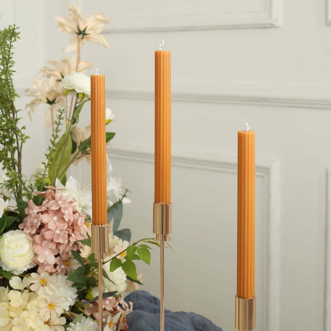 Three orange taper candles in elegant holders against a floral arrangement.