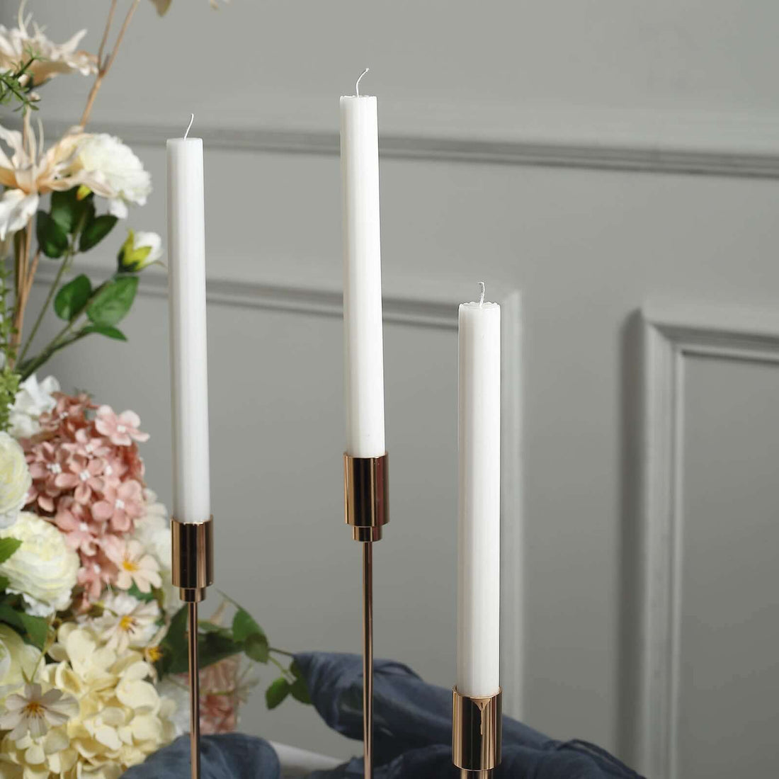 White candlesticks in elegant holders with floral backdrop.