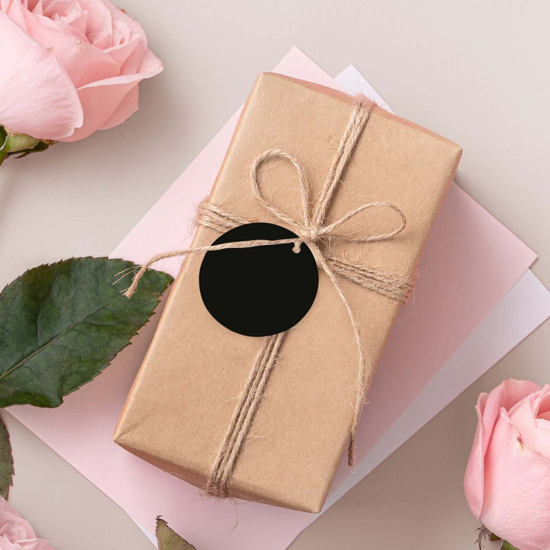 Gift box wrapped in brown paper with twine and a black tag, surrounded by pink roses.