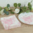 Pink floral napkins stacked beside a plate and greenery on a wooden surface.