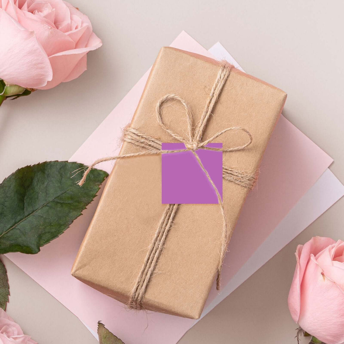 Gift box with twine and a purple tag, surrounded by pink roses and pastel papers.