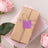 Gift box with twine and a purple tag, surrounded by pink roses and pastel papers.