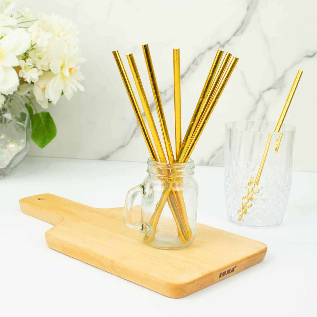 Gold striped paper straws in a jar on a wooden cutting board with flowers in the background.
