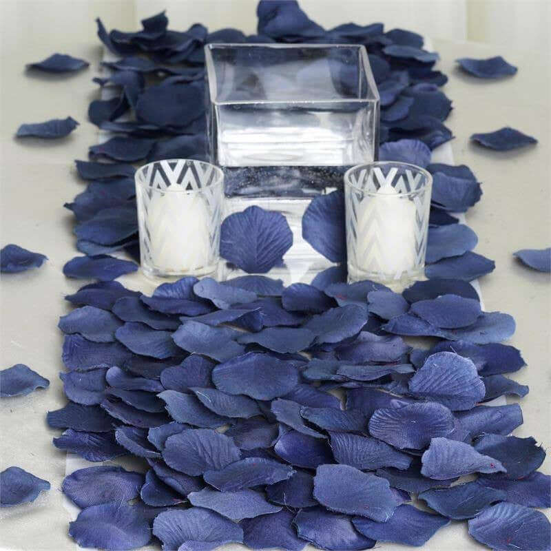 Navy blue rose petals scattered on a table with glass candle holders and a vase.