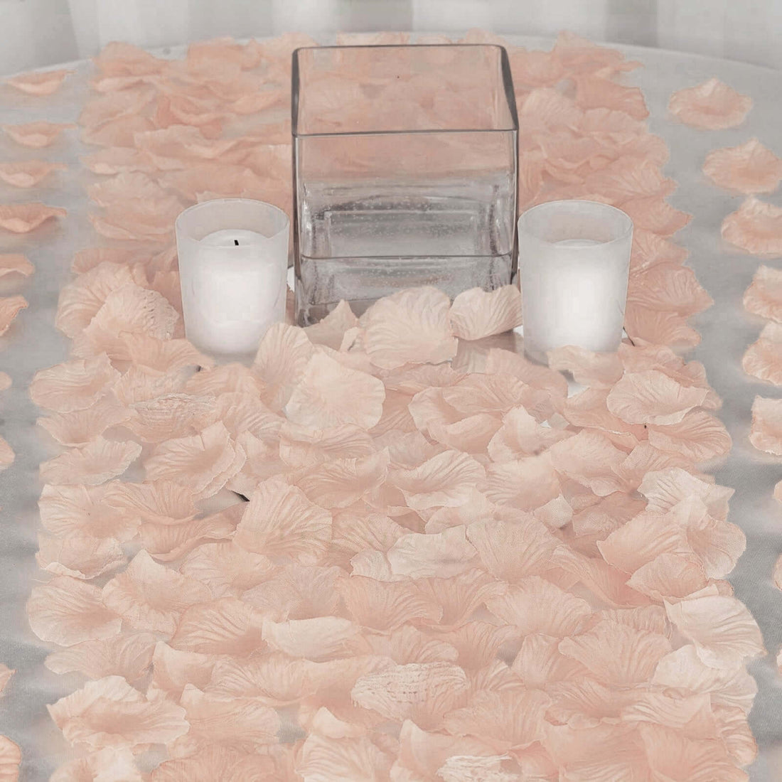 Pink rose petals scattered on a table with candles and a glass vase.