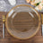 Gold beaded glass charger plate on wooden table with cutlery and flowers.