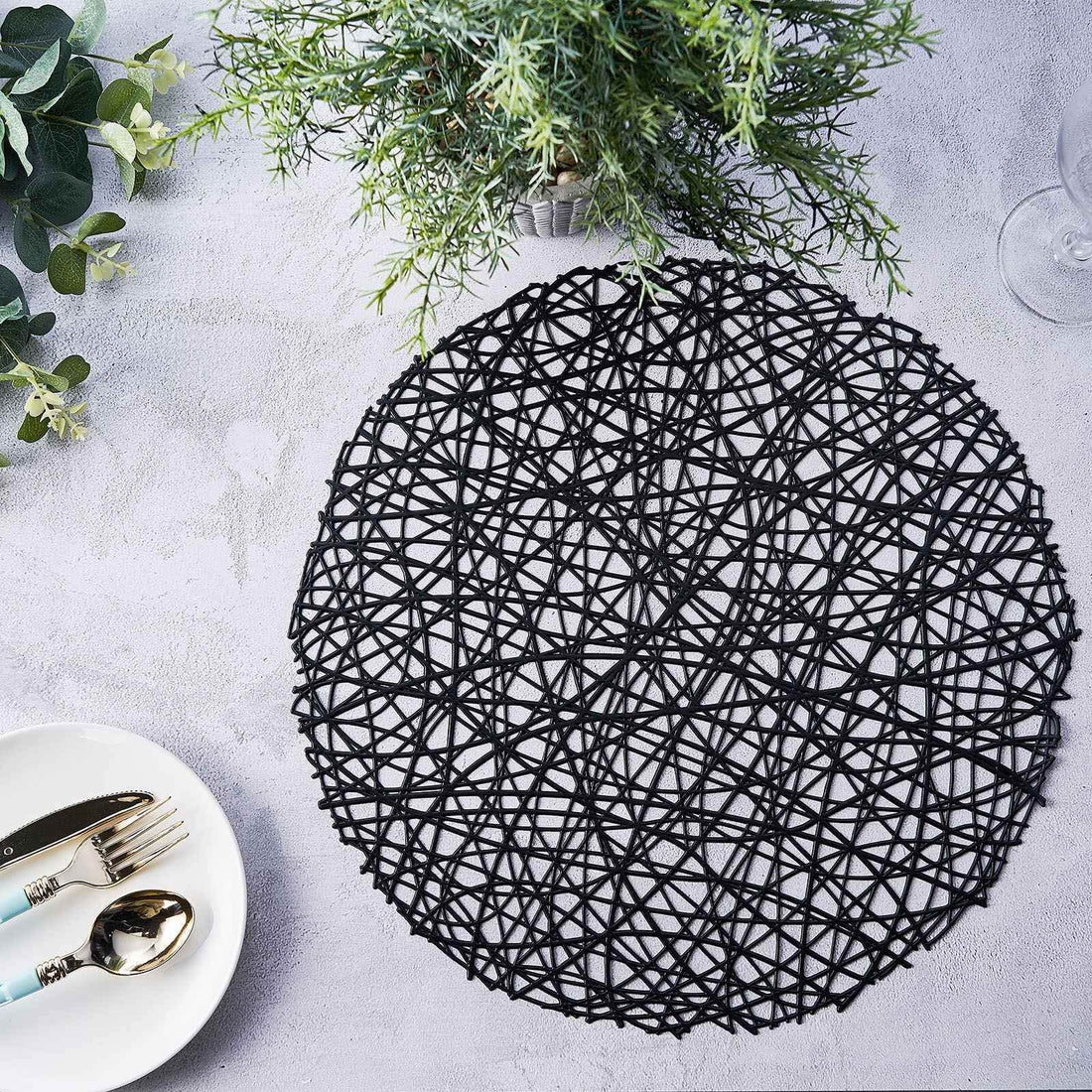 Black woven placemat on a textured surface with greenery and dining utensils.