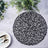 Black woven placemat on a textured surface with greenery and dining utensils.