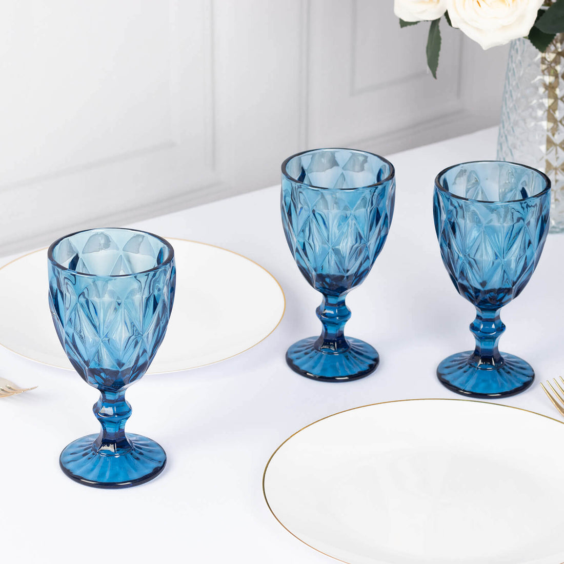 Set of three blue glass goblets on a table with white plates and floral decor.