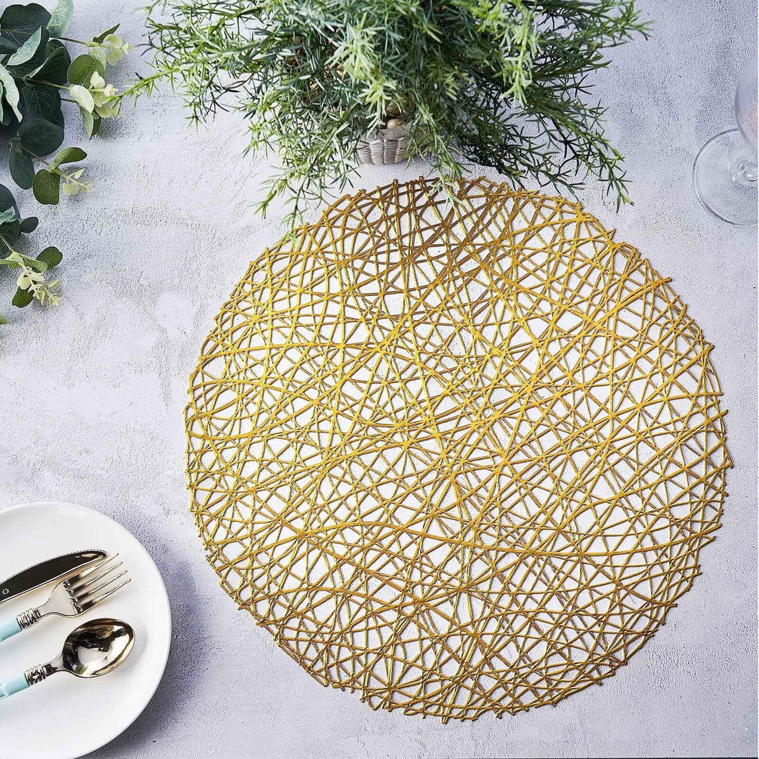 Gold woven placemat on a textured surface with greenery and cutlery.