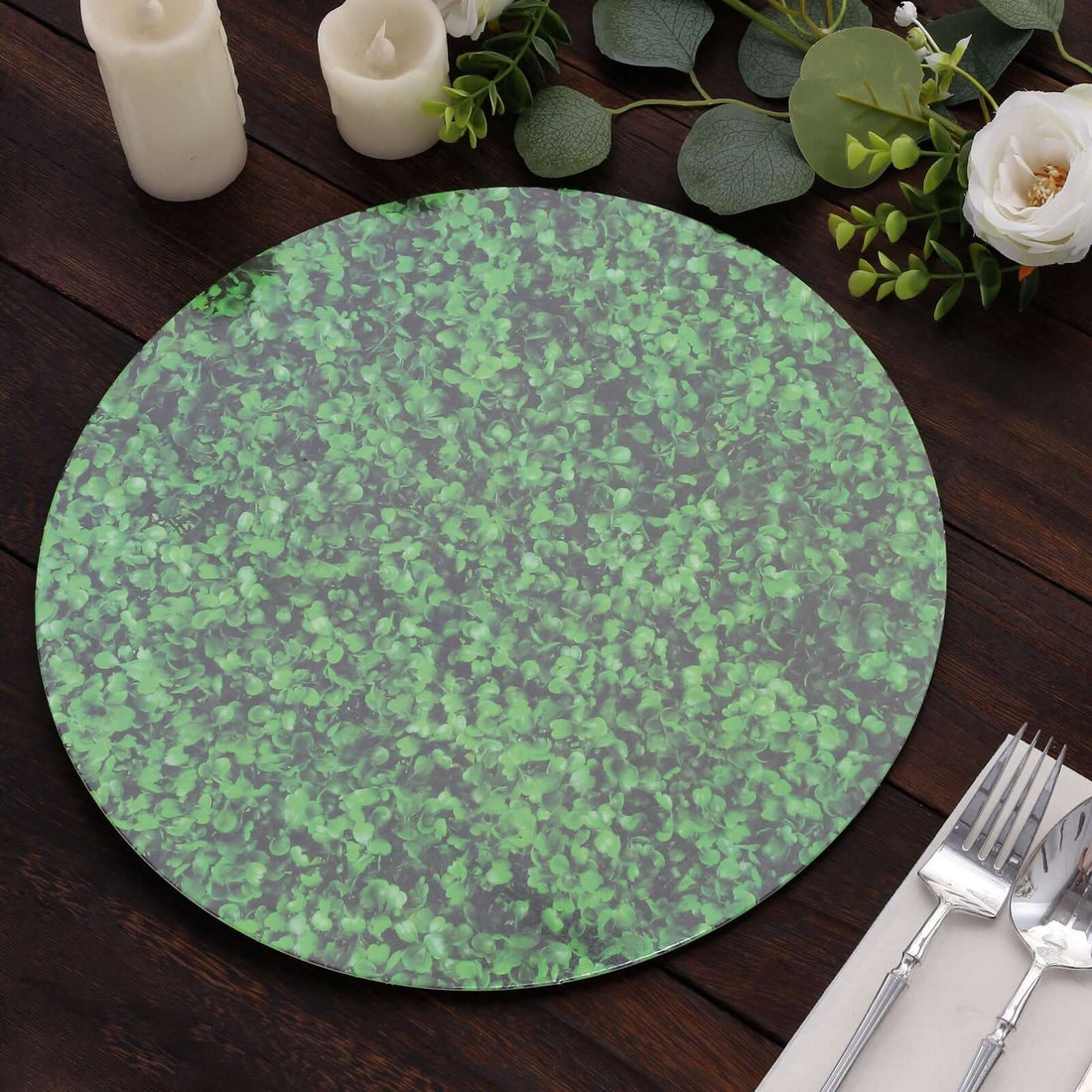 Green leaf-patterned round placemat on a wooden table with candles and flowers.