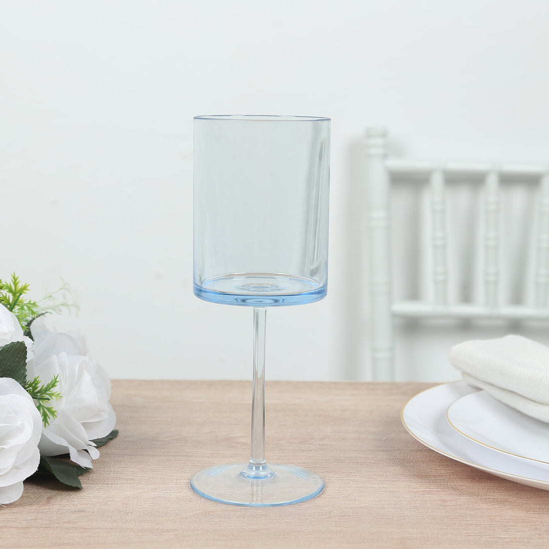 Blue stemmed glass on a wooden table with white flowers and plates.