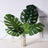 Artificial monstera leaves in a gold vase on a light wood surface.