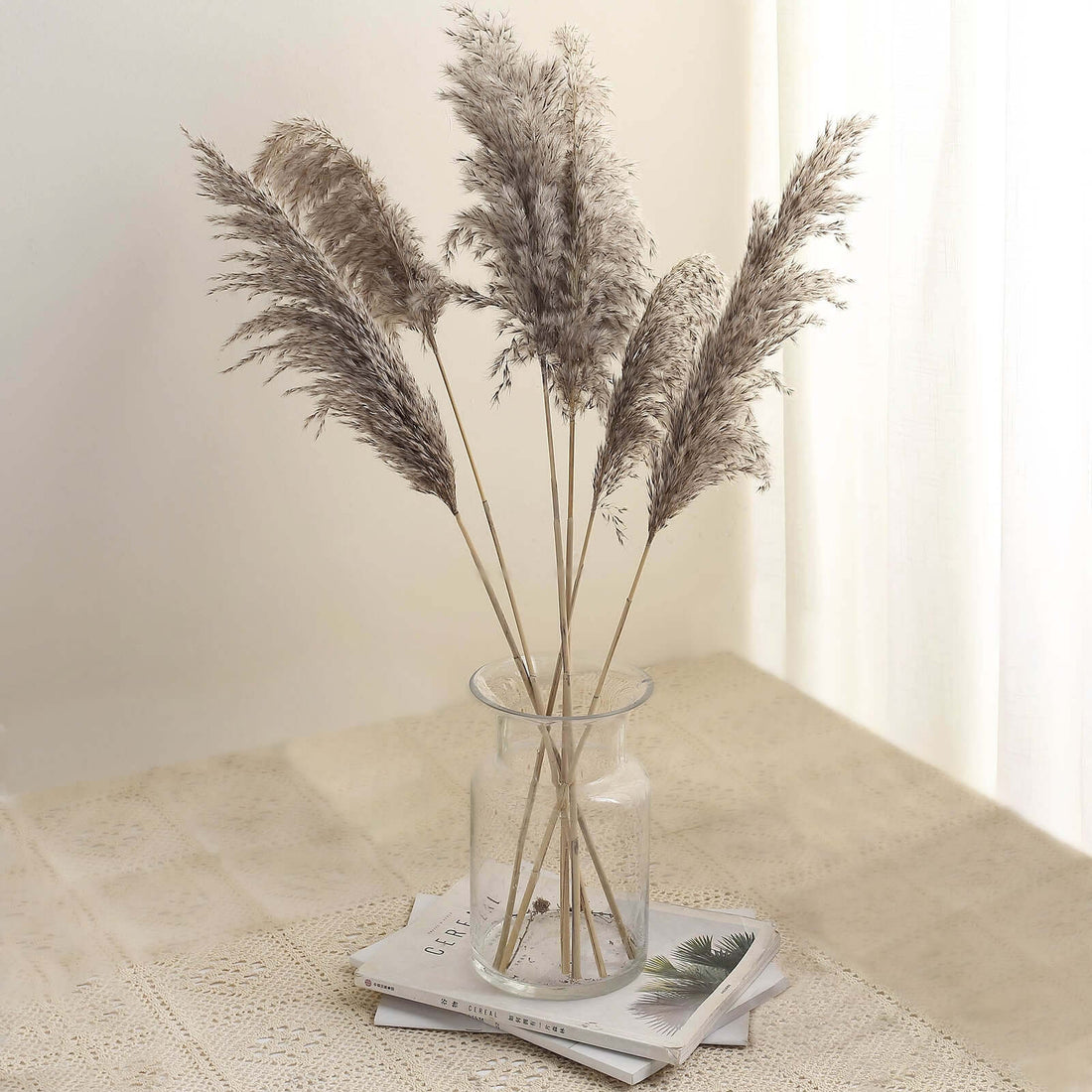 Dried pampas grass in a glass vase on a table with magazines.