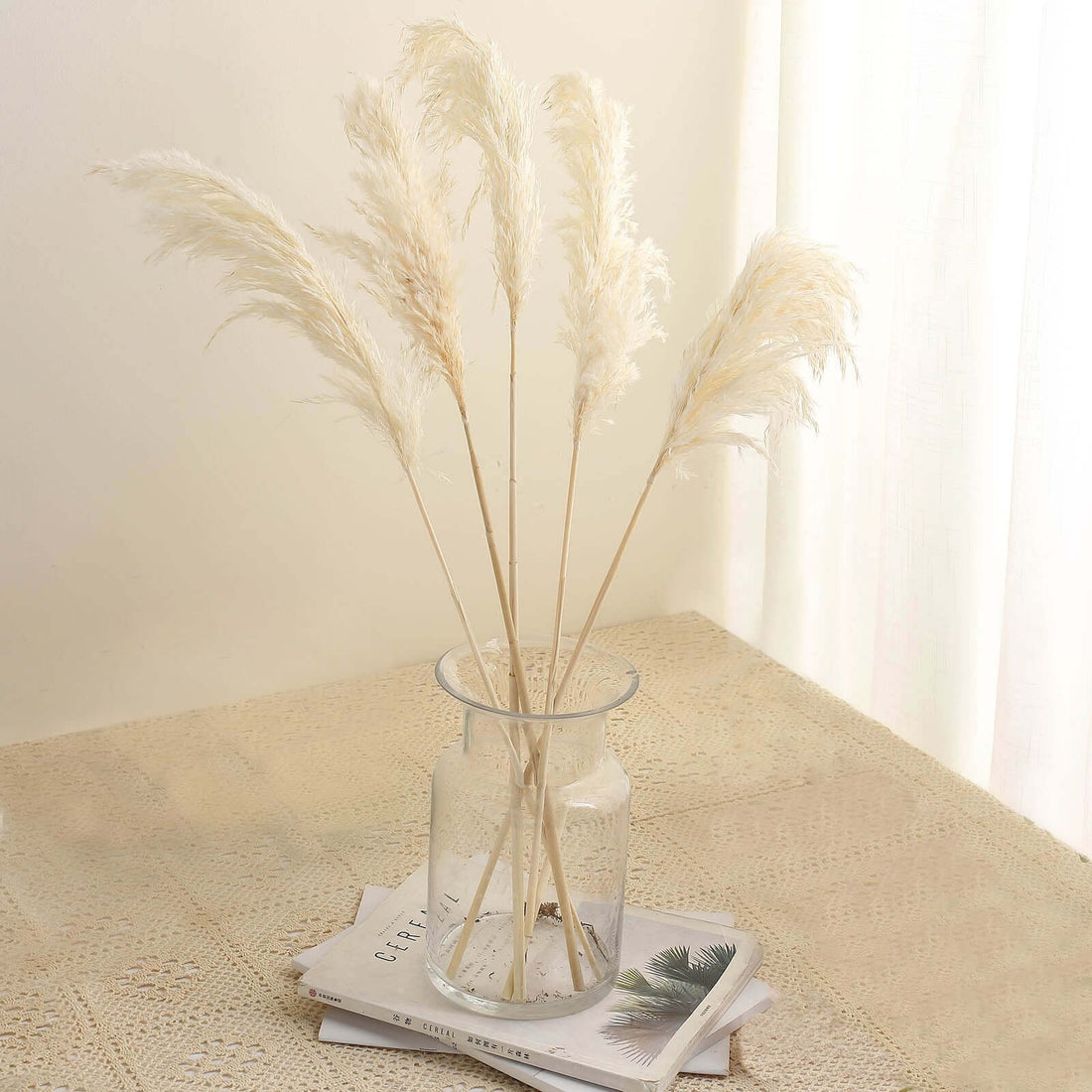 Dried pampas grass in a glass vase on a stack of magazines.