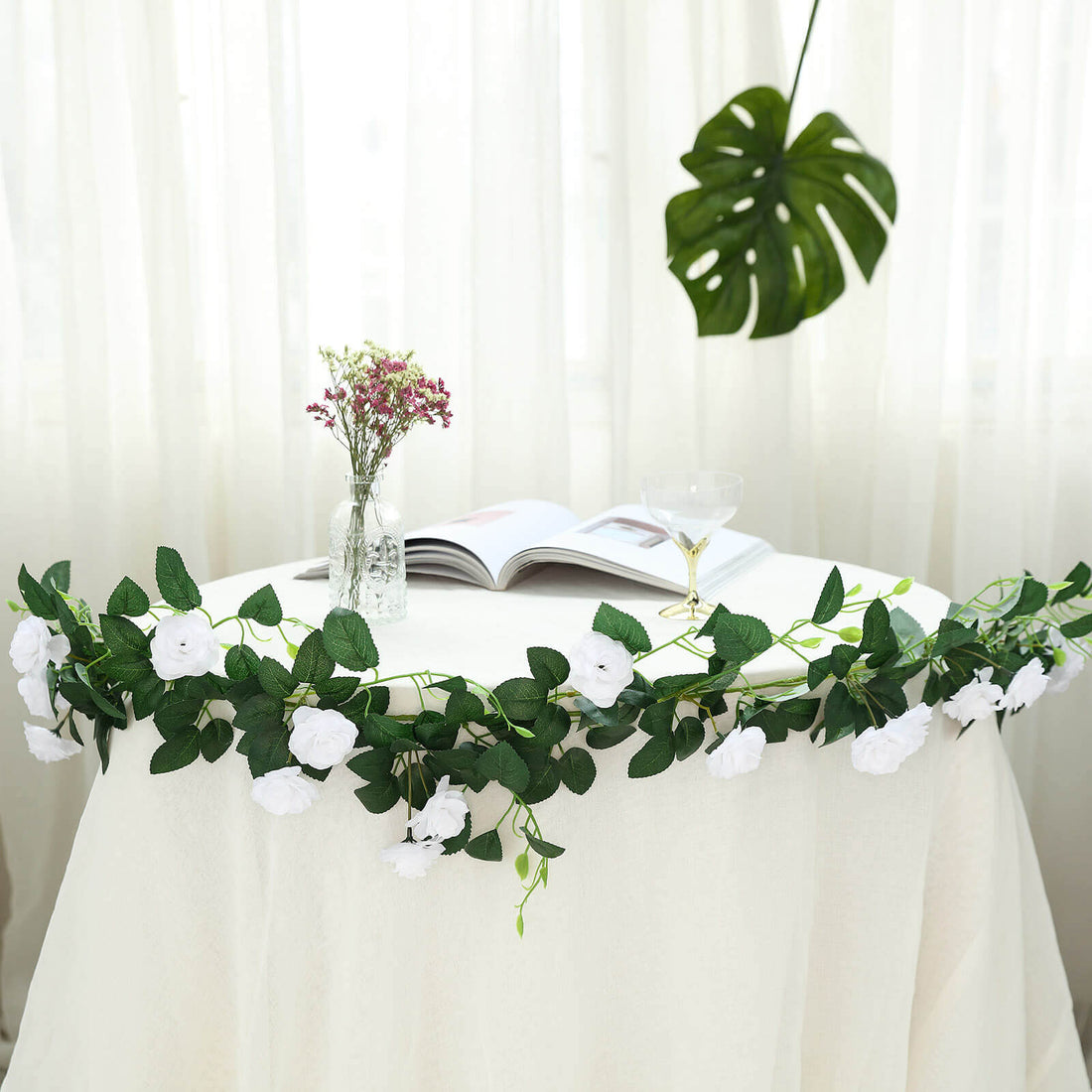 Artificial white rose garland on a table with a book and glass.