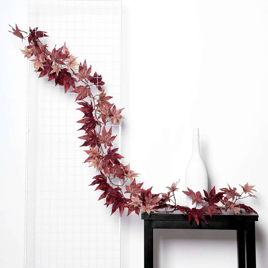Artificial red maple leaf garland on a black table next to a white vase.